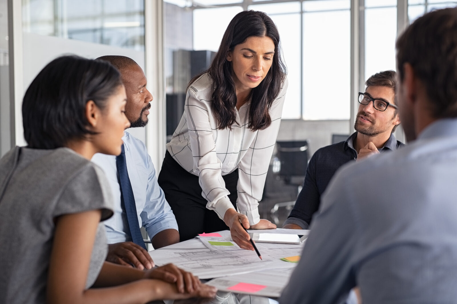 Woman with long brown hair standing over table of colleagues pointing at and presenting paperwork in a business discussion with buying committee.